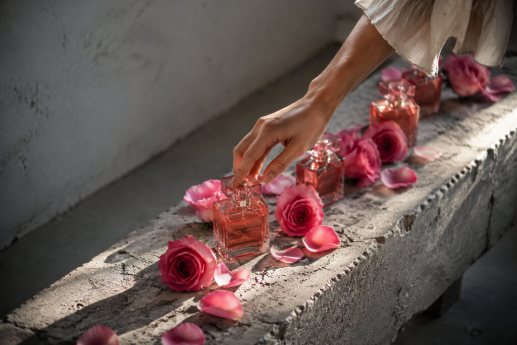 Une main effleure un flacon de parfum entouré de roses sur un banc en béton.