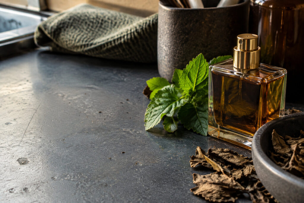 Flacon de parfum ambré entouré de feuilles fraîches et séchées de patchouli sur une table en pierre sombre.